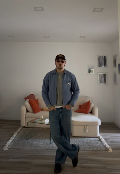 Hombre con gafas de sol, gorra, chaqueta azul y vaqueros, de pie con las manos en los bolsillos en una sala de estar minimalista con sofá beige y cojines naranjas.