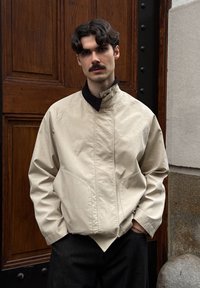 Veste légère beige avec un col montant, des poches latérales et des détails de couture visibles, associée à un pantalon sombre sur un fond en bois.