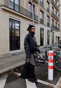 Chaqueta de cuero negra con collar alto, combinada con una camisa de cuadros atada a la cintura. Boina negra y bolso negro grande completan el conjunto.