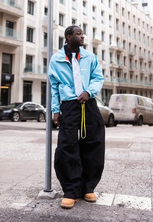 Hombre con chaqueta azul clara, corbata blanca con rayas, pantalones negros holgados con cordón amarillo y botas color marrón claro, de pie en una acera de la ciudad.