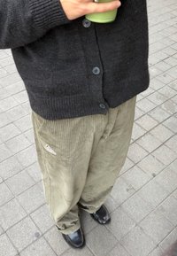 Zwarte gebreide cardigan met knopen, gecombineerd met olijfgroene ribfluwelen broek met verticale strepen en een wit logo. Zwarte schoenen zichtbaar.