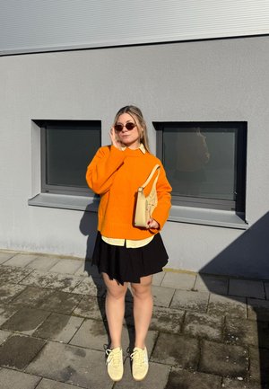 Mujer con suéter naranja, falda negra, camisa amarilla, bolso beige y gafas de sol, de pie sobre pavimento iluminado por el sol frente a una pared gris con dos ventanas.