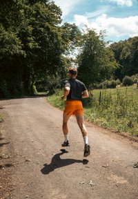 Coureur en chemise noire et shorts orange sur un chemin en gravier, entouré de verdure et d'arbres sous un ciel bleu dégagé.