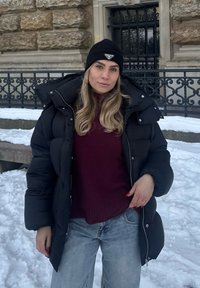 Femme portant un bonnet noir, une doudoune noire, un pull bordeaux et un jean, debout dans la neige devant un mur de pierre et une rampe en fer.