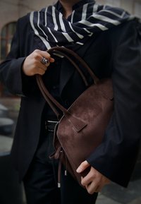 Smeđa torba od brušene kože s dva zaobljena ručka, zatvaranjem na vezanje i suptilnim detaljima šavova. Nošena uz crno odijelo i prugasti šal.