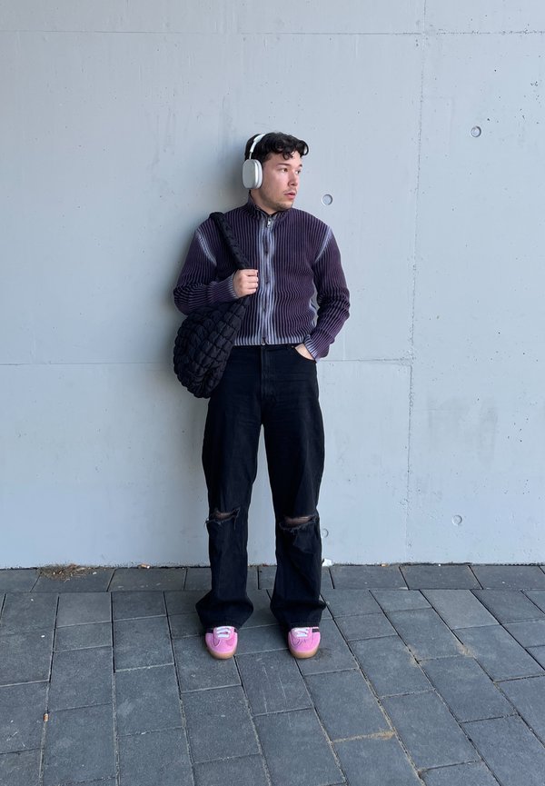 Modelo con un suéter morado de canalé con cremallera, jeans negros rasgados, calzado rosa y un bolso negro de mimbre oversized. Lleva puestos auriculares blancos.