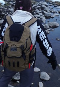 Personne portant un sac à dos beige et des gants noirs marchant sur des rochers près d'un ruisseau peu profond.