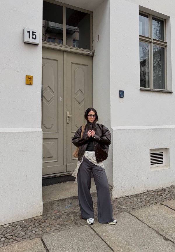 Žena v okuliaroch s hnedou bundou, sukňou s čipkovou lemovkou cez široké sivé nohavice a teniskami stojí na kamennej chodníku pri béžových dverách.