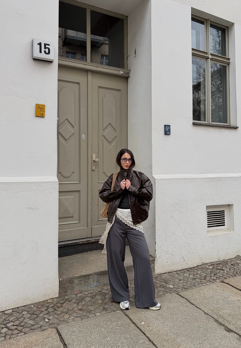 Femme avec lunettes, veste marron, jupe bordée de dentelle par-dessus un pantalon gris large et des baskets, debout sur un trottoir en pierre près d'une porte beige.