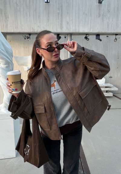 Chaqueta bomber de cuero marrón con un ajuste relajado, que cuenta con grandes bolsillos delanteros y un cuello alto. Sudadera gris debajo y gafas de sol.