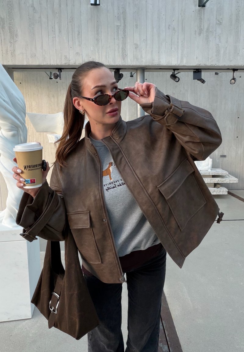 Veste aviateur en cuir marron avec une coupe décontractée, dotée de grandes poches avant et d'un col haut. Sweat-shirt gris en dessous et lunettes de soleil.