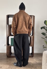 Personne portant une veste marron, un bonnet noir et un pantalon noir ample, debout à l'intérieur face à une étagère en bois avec des livres et une plante à proximité.