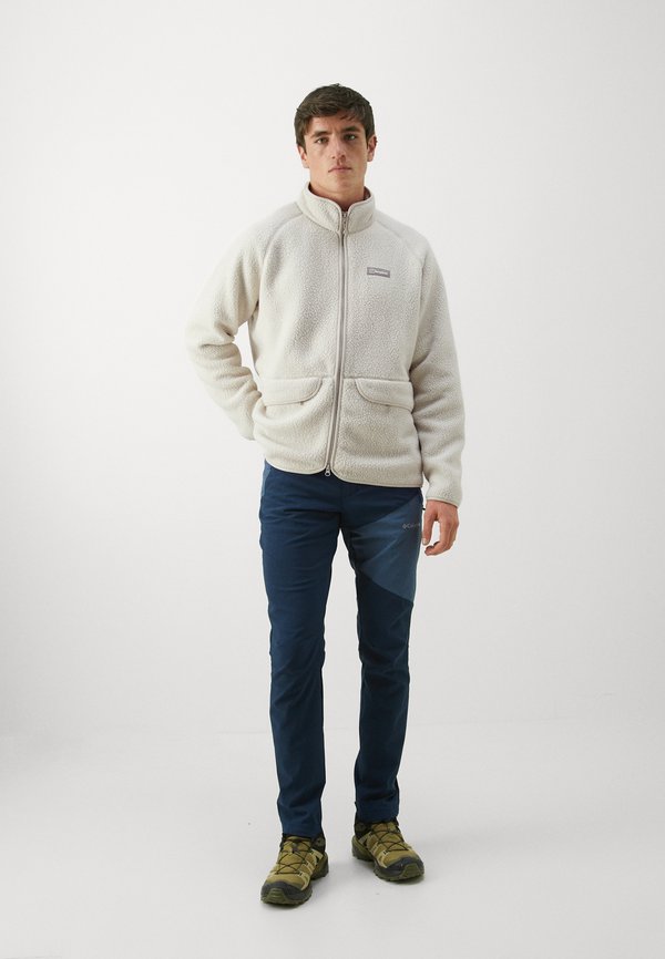 Valgebeež fliisjakki kõrge kraega, luku ees ja kahe esitaskuga. Seljas tumesinised püksid ja rohelised matkajalatsid.