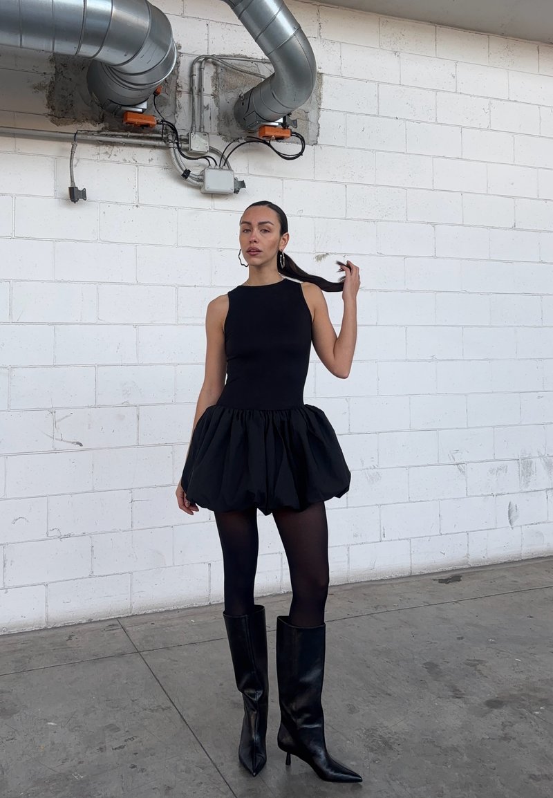 Donna che sistema i capelli, indossando un abito nero senza maniche con gonna gonfia, collant neri e stivali neri fino al ginocchio in uno spazio industriale.