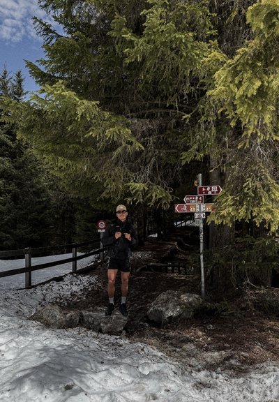 Hombre con mochila de pie en un sendero forestal nevado junto a varias señales direccionales de senderismo y ciclismo bajo grandes árboles perennes.