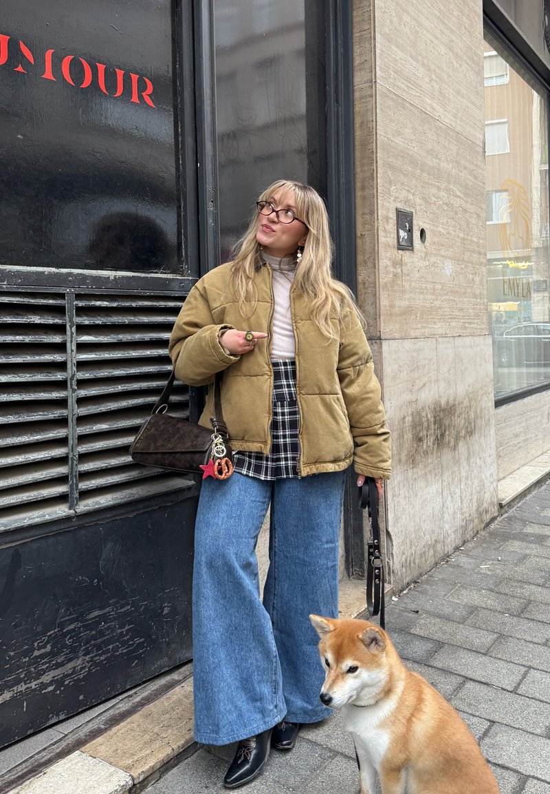Femme portant une veste marron, une chemise à carreaux, un jean large et des lunettes, tenant une laisse de chien tout en se tenant sur un trottoir en ville avec un chien beige et blanc.