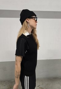 Sort strik sweater med en hvid krave, sort hue og solbriller. Langt hår stylet nedad, med sølv øreringe og synlige tatoveringer.