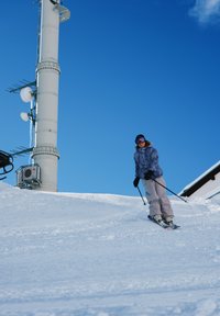 Skis avec fixations colorées sur une pente enneigée. Un skieur porte une veste bleu clair, un pantalon beige et des gants noirs sous un ciel bleu clair.