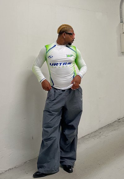 Camisa deportiva de manga larga en blanco con detalles en verde y azul, combinada con pantalones grises holgados; ambos presentan un tejido transpirable. Calzado negro.
