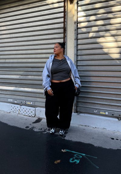 Top corto gris, sudadera oversized gris, pantalones holgados negros y zapatos negros con acentos grises, de pie frente a un fondo de persiana metálica.