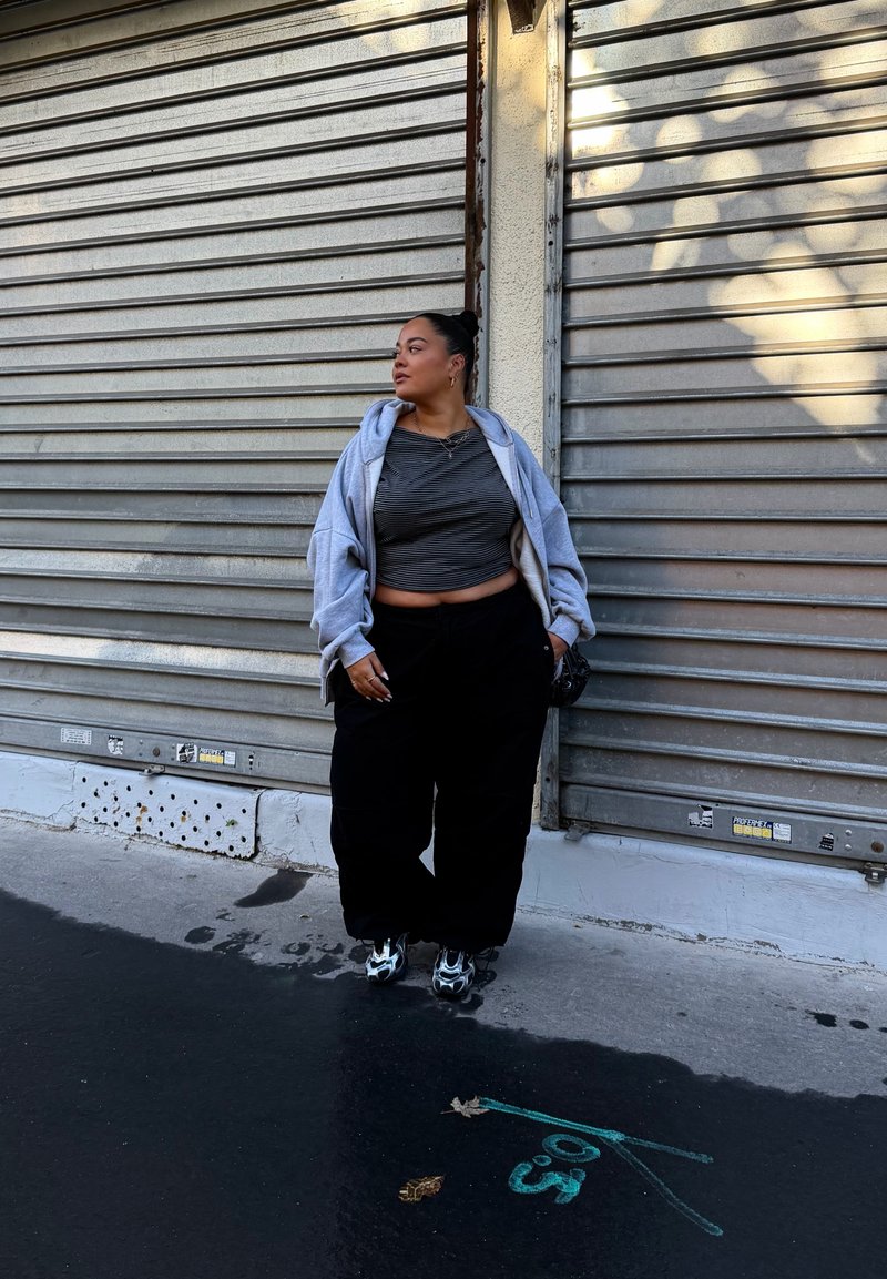 Šedý krátky top, šedá voľná mikina, čierne voľné nohavice a čierne topánky so šedými akcentmi, stojí proti pozadiu z kovových roliet.