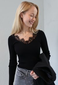 Mustast nööbitud pikad varrukad ja pitsist V-kaelusega topp. Kombineeritud sädelevate kaunistustega halli teksadega. Käes hoiab musta jope.