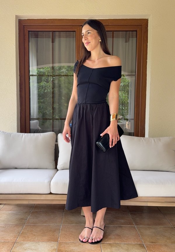 Frau in schwarzem schulterfreiem Kleid, die eine schwarze Clutch hält und ein goldenes Armreif trägt, steht drinnen auf gefliestem Boden nahe einem weißen Sofa.