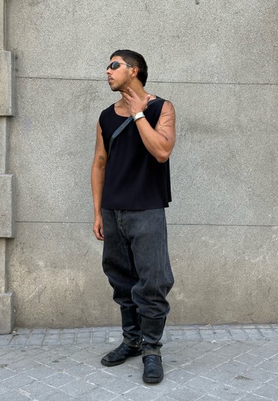 Top negro de canalé, pantalones de mezclilla oscuros y holgados, y botas negras hasta la rodilla. Los accesorios incluyen gafas de sol y un reloj. Fondo gris texturizado.