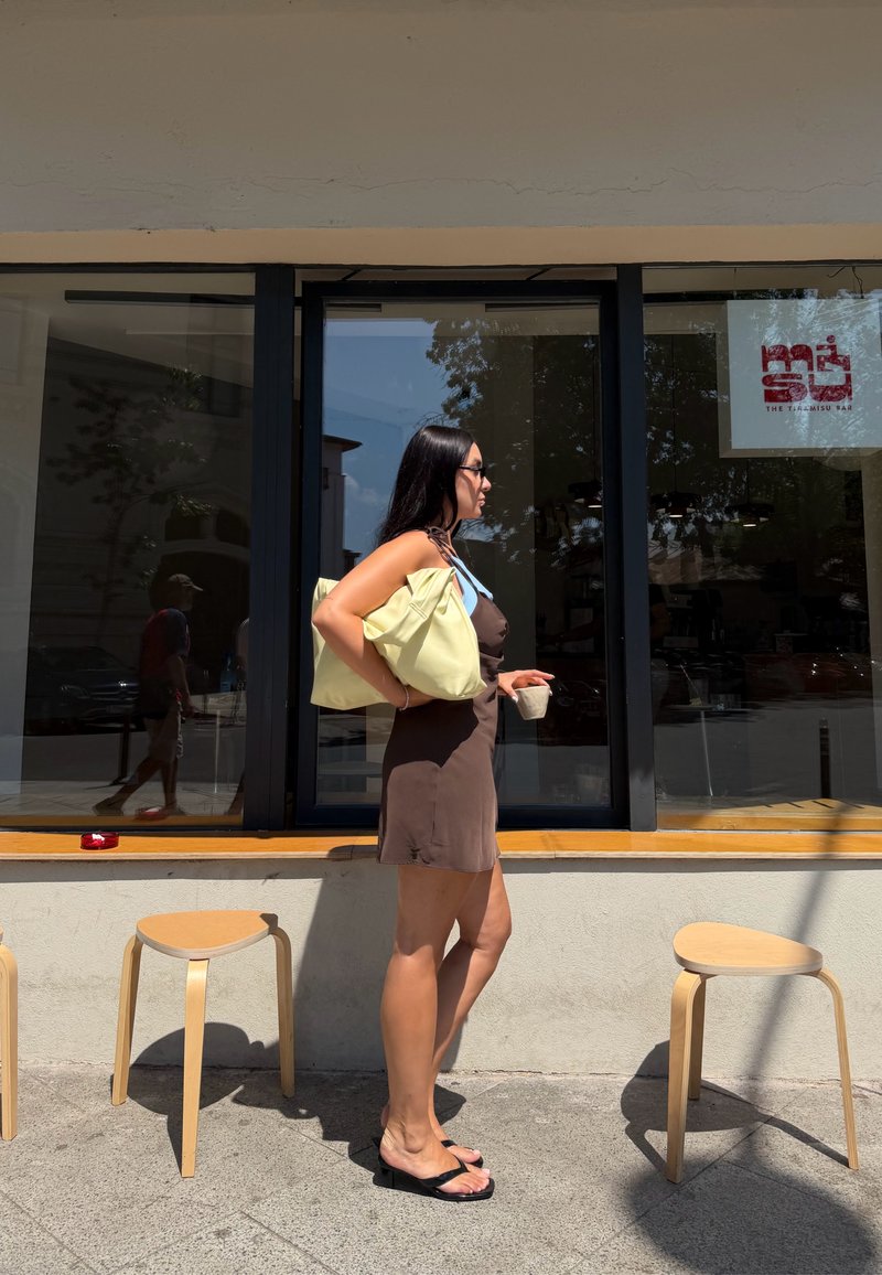 Femme en robe marron tenant un sac jaune et une tasse de café, debout entre deux tabourets en bois devant un café avec de grandes fenêtres.