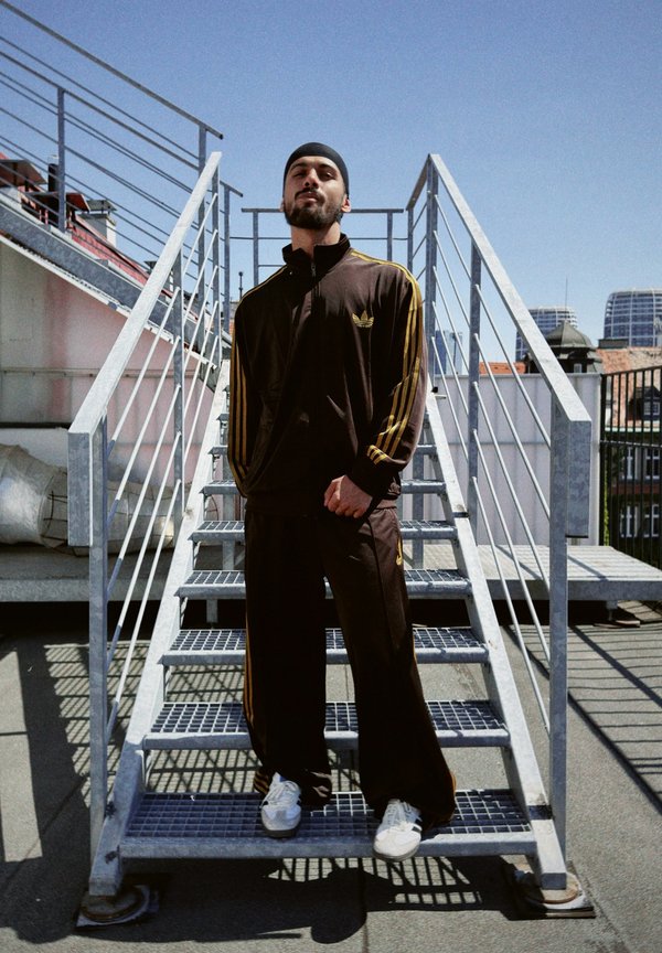 Completo sportivo marrone con strisce gialle, sneakers bianche, in piedi su scale di metallo. Cielo limpido e edifici della città sullo sfondo.