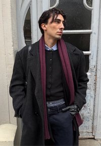 Abrigo largo de lana negra, suéter oscuro con botones, camisa a rayas, bufanda granate, pantalones azul marino, guantes de cuero, de pie junto a una puerta texturizada.