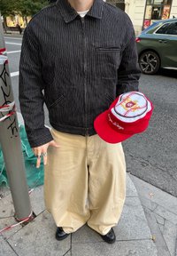 Fekete vékony csíkos zakót és széles bézs nadrágot viselő személy, aki egy piros-fehér baseball stílusú sapkát tart egy városi járdán.