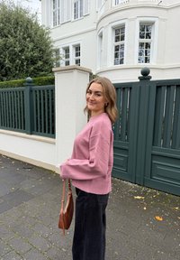 Różowy sweter z prążkowanym dołem, zestawiony z czarnymi szerokimi spodniami. Trzyma małą brązową torebkę. W tle biały budynek i zielone ogrodzenie.