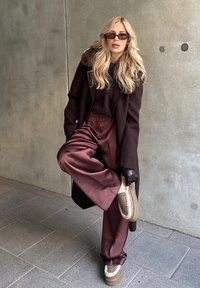 Manteau marron avec col en fourrure, pull sombre, pantalon en satin bordeaux et chaussures marron sans lacets avec des accents clairs. Des lunettes de soleil élégantes complètent le look.