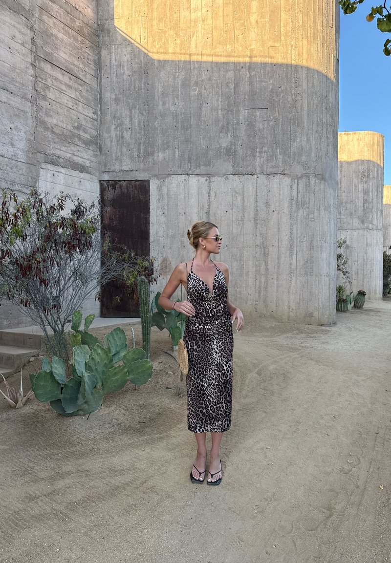 Femme en robe à imprimé léopard et lunettes de soleil, debout sur un sol sablonneux près de grands cylindres en béton et de plantes de cactus.