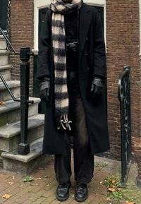 Μαύρο παλτό στρωμένο πάνω από ένα μαύρο πουλόβερ, συνδυασμένο με φαρδιά μαύρα παντελόνια. Περιλαμβάνει ένα καρό κασκόλ και μαύρα δερμάτινα γάντια.