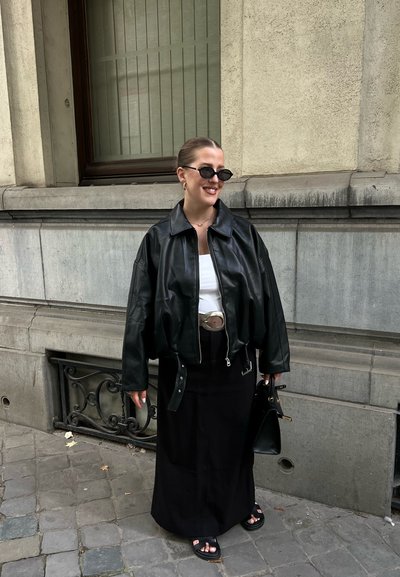 Mujer vestida con chaqueta de cuero negra, blusa blanca, falda negra, sandalias negras, gafas de sol y llevando un bolso negro, de pie sobre un pavimento de piedra.