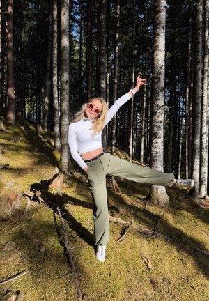 Mujer joven con gafas de sol, llevando un top corto blanco y pantalones verdes, equilibrándose en una pierna con el brazo levantado en un bosque iluminado por el sol.