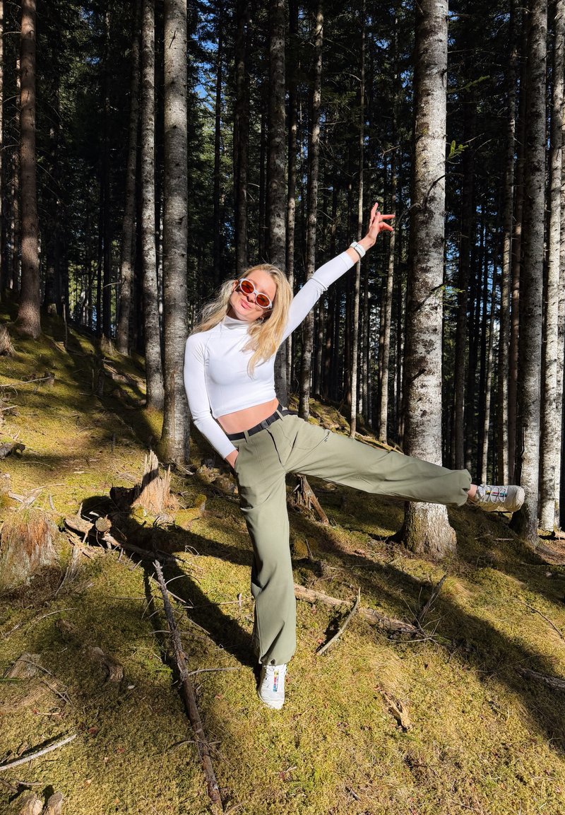 Νεαρή γυναίκα με γυαλιά ηλίου, φοράει λευκό crop top και πράσινο παντελόνι, ισορροπεί στο ένα πόδι με το χέρι σηκωμένο σε φωτεινό δάσος.