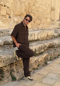 Cămășă marron cu mânecă scurtă, cu două buzunare pe piept și pantaloni asortați. Tenisi casual cu un design în nuanțe de marron și crem. Fond de piatră.