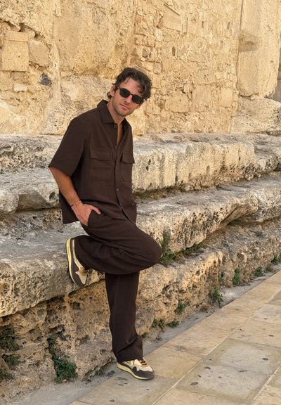 Camisa marrón de manga corta con dos bolsillos en el pecho y pantalones a juego. Las zapatillas deportivas informales tienen un diseño marrón y crema. Fondo de piedra.