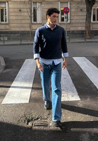 Άνδρας με σκούρα σγουρά μαλλιά, φοράει μπλε πουλόβερ και τζιν, περπατώντας σε διάβαση πεζών σε αστική οδό κατά τη διάρκεια της ημέρας.