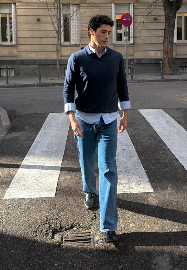 Mann med mørkt krøllete hår iført marineblå genser og jeans som går over fotgjengerovergang på bygate i dagslys.