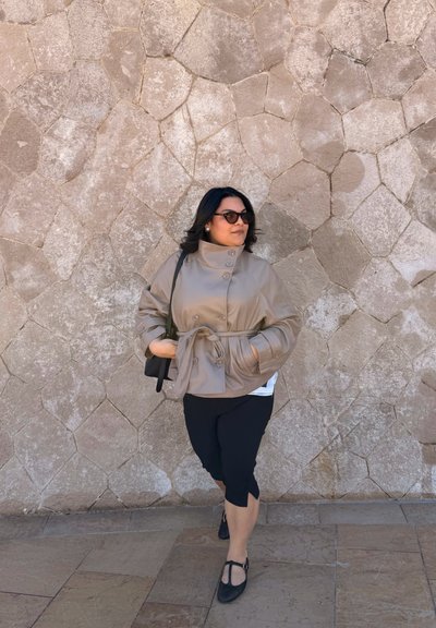 Mujer con chaqueta beige con cinturón, pantalones negros recortados y gafas de sol caminando frente a una pared de piedra tostada con patrón geométrico.