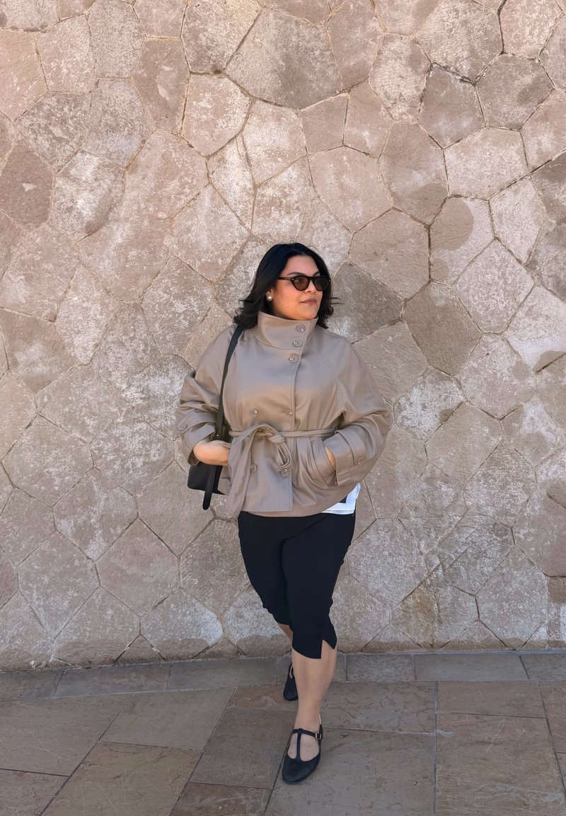 Moteris dėvi smėlio spalvos diržu aprištą švarką, juodus trumpintus kelnes ir akinius nuo saulės, žingsniuoja prie šviesiai rudos akmeninės sienos su geometriniu raštu.