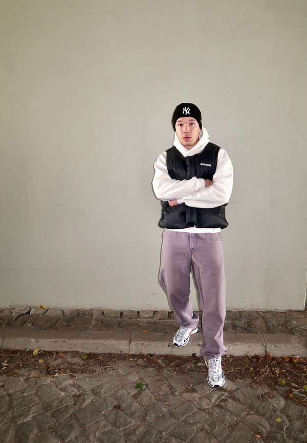 Black puffer vest worn over a white hoodie, paired with light grey trousers and silver sneakers. A black beanie featuring a logo completes the outfit. Background: smooth grey wall.