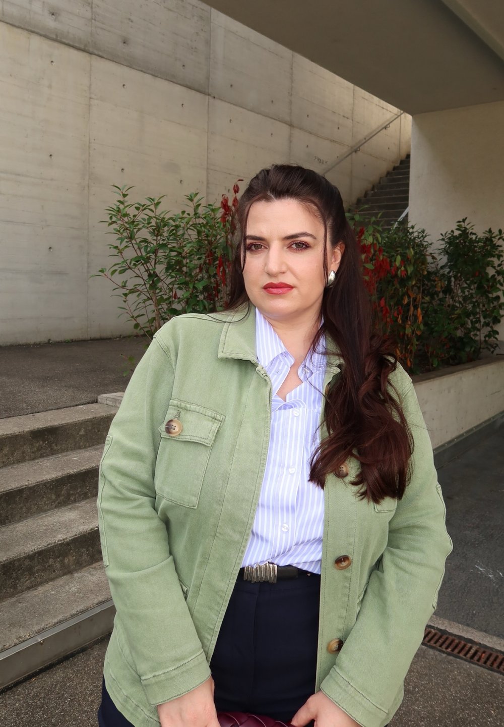 Vrouw met lang bruin haar, gekleed in een groene jas en gestreept shirt, staat buiten bij betonnen trappen en planten onder een poort.