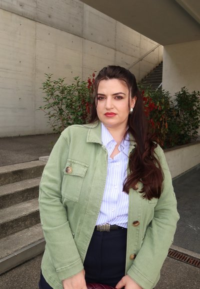 Mujer con cabello largo castaño que lleva una chaqueta verde y una camisa de rayas, de pie al aire libre cerca de unas escaleras de concreto y plantas bajo un arco.