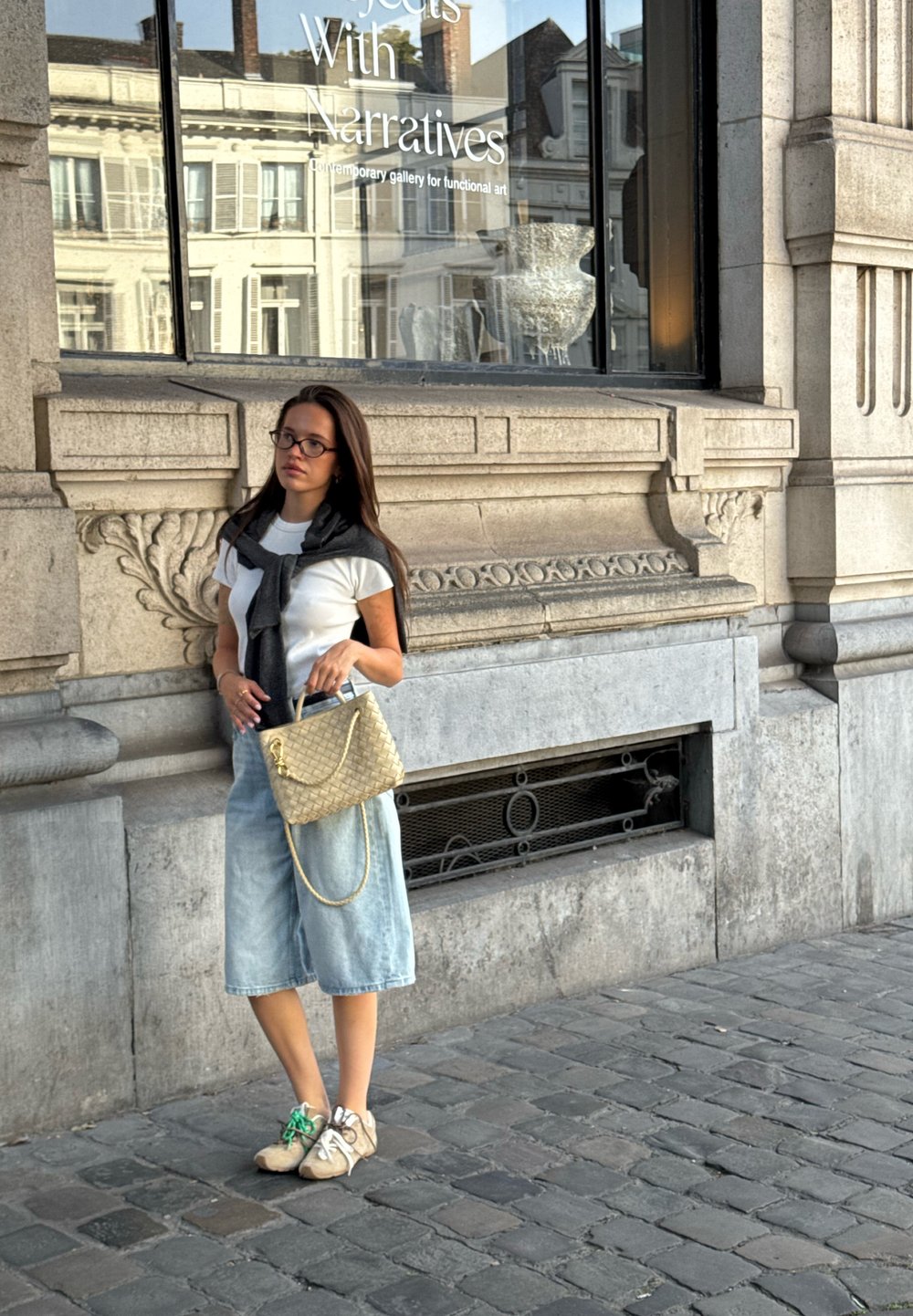 Denim-Culottes, weißes T-Shirt, dunkler Pullover über die Schultern gelegt, beiger Woven-Handtasche und beigefarbene Sneakers mit grünen Akzenten. Kopfsteinpflasterstraße im Hintergrund.