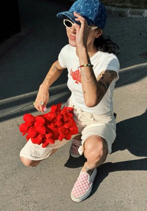Camiseta blanca con logo rojo, pantalones cortos beiges y zapatillas de cuadros rosas. Sosteniendo un ramo de flores de tela rojas. Gorra de mezclilla azul.